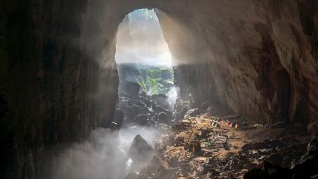 View of an opening to Son Doong cave in Vietnam. Light filters into the cave and we see people camping on a ledge.